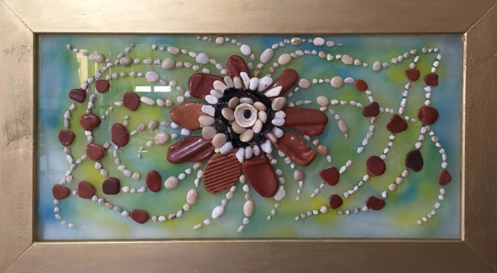 Composición floral realizada con piedras naturales sobre plexiglás. Una gran flor central de tonos rojizos se abre sobre un fondo verde y azul, rodeada por delicados trazos curvos formados por pequeñas piedras blancas y rojizas.