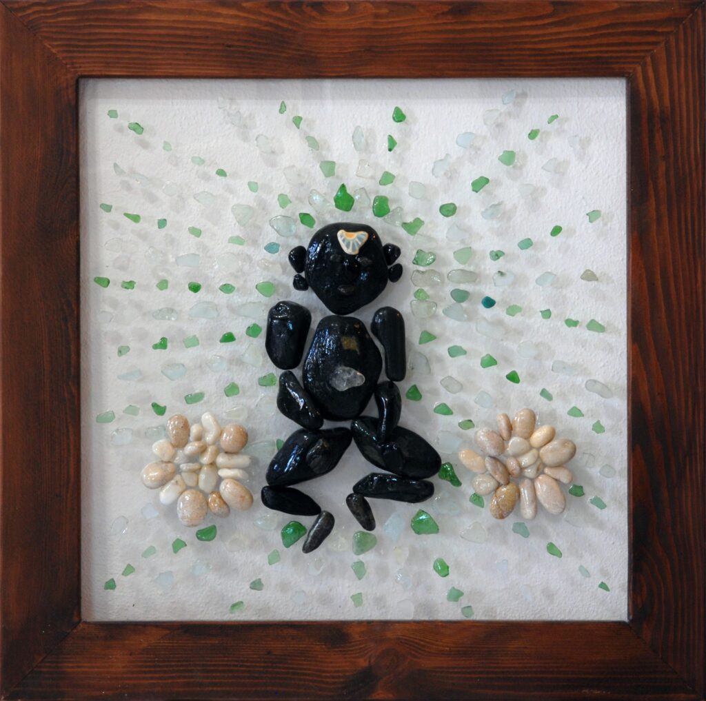 Figura inspirada en Buda realizada con piedras negras sobre plexiglás, rodeada por fragmentos de vidrio verde y transparente que irradian hacia el exterior.