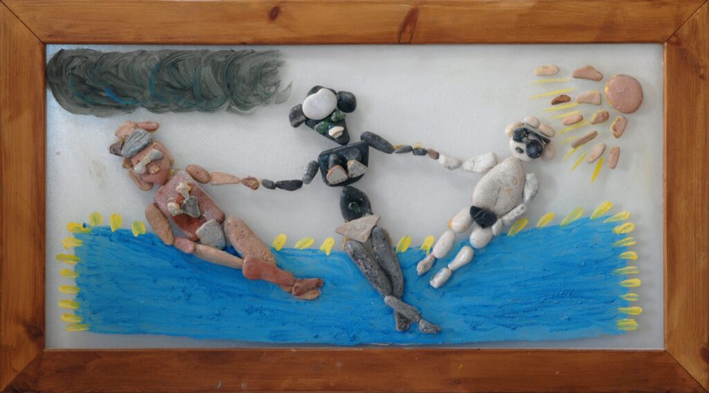 Scena realizzata con pietre marine su plexiglas che rappresenta una famiglia in spiaggia, con i genitori che tengono per mano il proprio bambino accanto al mare.