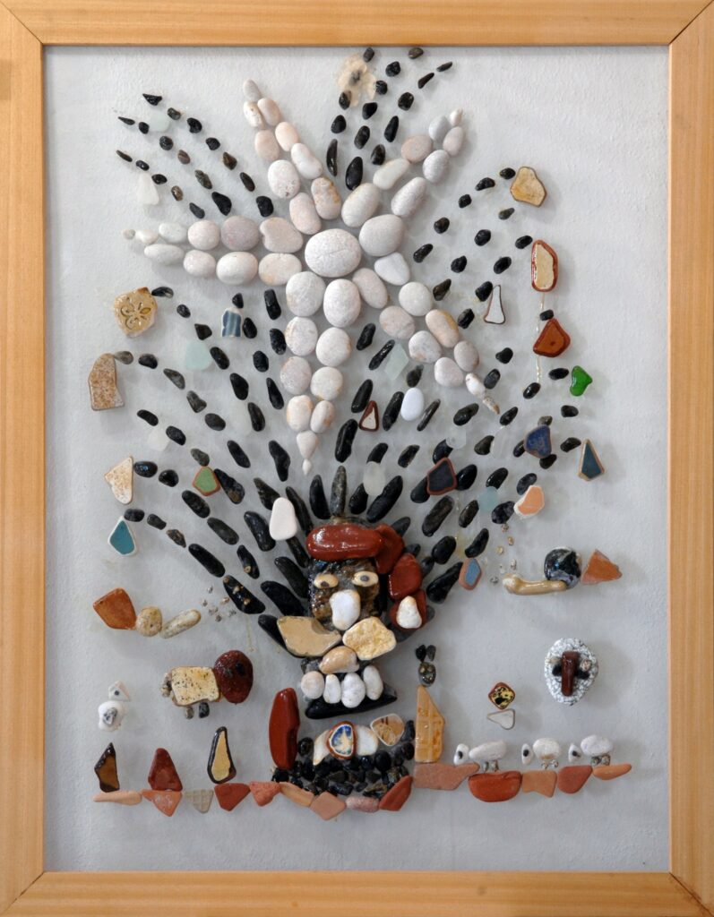 Composición realizada con piedras marinas, fragmentos de cerámica y vidrio sobre plexiglás que representa un nacimiento navideño con una estrella brillante, animales, figuras humanas y elementos simbólicos del pesebre.