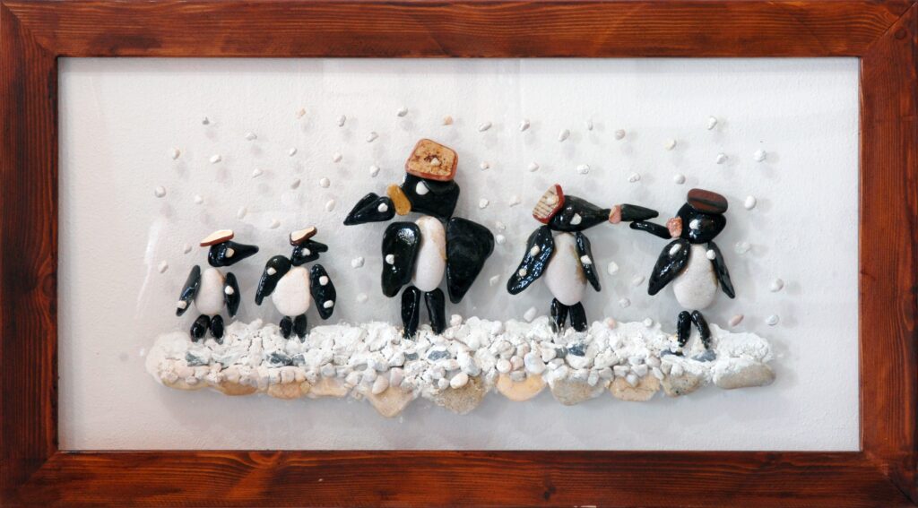 Familia de pingüinos realizada con piedras naturales sobre plexiglás, caminando sobre la nieve mientras caen suaves copos en un paisaje invernal.