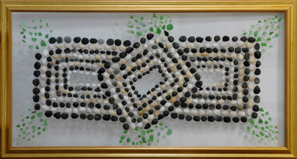 Composición geométrica realizada sobre vidrio con piedras blancas, negras y grises formando una estructura simétrica entrelazada, rodeada por pequeños fragmentos de vidrio verde en los extremos.